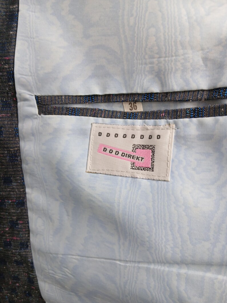May include: Close-up of a jacket's interior, showing a light blue patterned lining. The jacket's outer fabric is dark and textured, with blue and pink flecks. A tag reads "DDD DIREKT" and the size is 36.