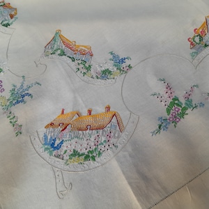 May include: White linen tablecloth with embroidered houses and flowers. The houses have yellow roofs and red brick walls. The flowers are in shades of pink, blue, and yellow.