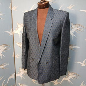 May include: A dark blue double-breasted blazer with a small square pattern. The jacket has two rows of buttons and a notched lapel. The blazer is displayed on a mannequin, highlighting its tailored fit and classic design.