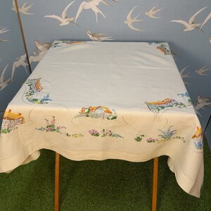 May include: A white linen tablecloth with an embroidered design of a cottage and flowers. The tablecloth is on a wooden table and has a scalloped edge.
