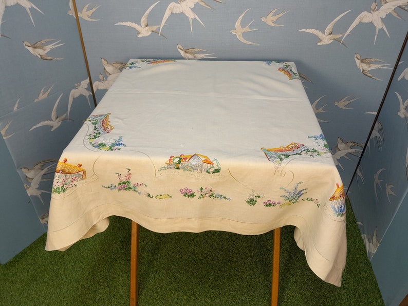 May include: A white linen tablecloth with an embroidered design of a cottage and flowers. The tablecloth is on a wooden table and has a scalloped edge.