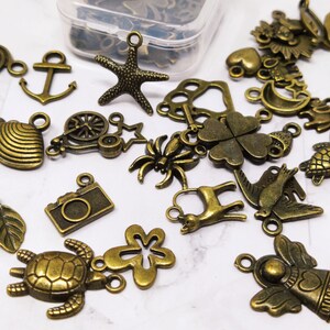 May include: Assortment of bronze-colored metal charms with various designs, including a camera, a turtle, a flower, a bird, a deer, a spider, a clover, a crescent moon, a star, a shell, a wheel, a leaf, an anchor, a spiral, and an angel.