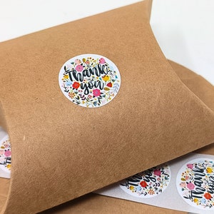 Pu&ograve; includere: Un rotolo di adesivi rotondi di ringraziamento con un design floreale e il testo "Grazie" in lettere nere. Gli adesivi sono su uno sfondo di carta marrone.
