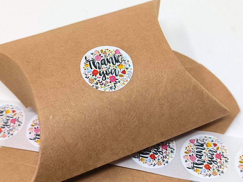 Pu&ograve; includere: Un rotolo di adesivi rotondi di ringraziamento con un design floreale e il testo "Grazie" in lettere nere. Gli adesivi sono su uno sfondo di carta marrone.