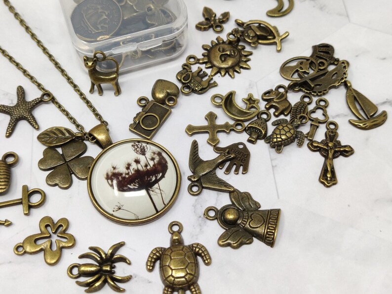 May include: A collection of bronze-colored charms and pendants, including a four-leaf clover, a bird, a cross, a star, a moon, an angel, a spider, a turtle, and a floral design. The charms are arranged on a white surface.