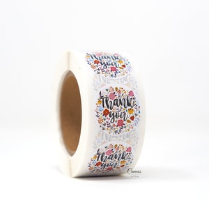 Pu&ograve; includere: Rotolo di adesivi rotondi con un design floreale e il testo "Grazie" in lettere nere.