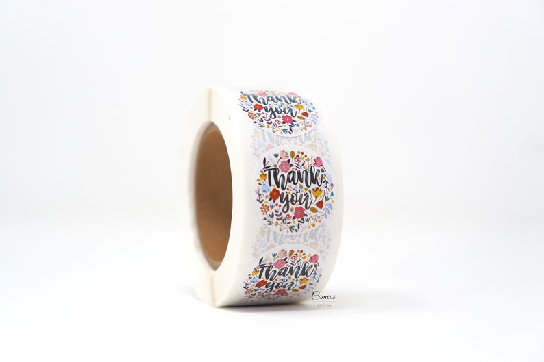 Pu&ograve; includere: Rotolo di adesivi rotondi con un design floreale e il testo "Grazie" in lettere nere.