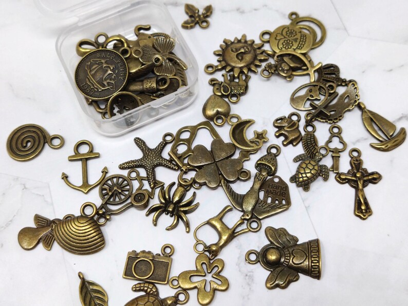 May include: A collection of antique gold-toned metal charms, including a star, a clover, a sun, a heart, a turtle, a fish, a spider, a camera, a leaf, a spiral, an anchor, a wheel, a bird, a crown, a skull, a bell, a hand-made tag, and a religious cross.
