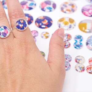 Puede incluir: Tres anillos de plata con inserciones de vidrio de patrones coloridos. Los anillos est&aacute;n en una mano, y hay muchos m&aacute;s insertos de vidrio coloridos en el fondo.