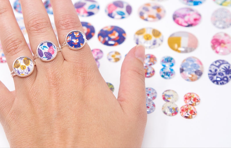Puede incluir: Tres anillos de plata con inserciones de vidrio de patrones coloridos. Los anillos est&aacute;n en una mano, y hay muchos m&aacute;s insertos de vidrio coloridos en el fondo.