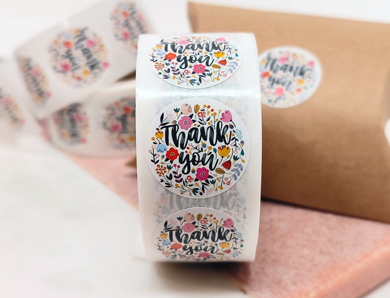 Pu&ograve; includere: Rotolo di adesivi rotondi con un design floreale e il testo "Grazie". Gli adesivi sono bianchi con fiori e foglie colorati.