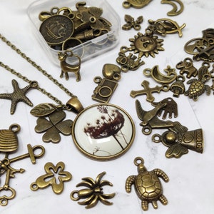 May include: A collection of bronze-toned metal charms and pendants, including a four-leaf clover, a starfish, a camera, a cross, a turtle, a spider, a bicycle, a bird, and a dandelion. The charms are arranged on a white surface with a necklace featuring a dandelion charm.