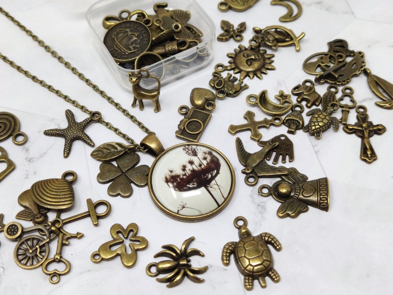 May include: A collection of bronze-toned metal charms and pendants, including a four-leaf clover, a starfish, a camera, a cross, a turtle, a spider, a bicycle, a bird, and a dandelion. The charms are arranged on a white surface with a necklace featuring a dandelion charm.