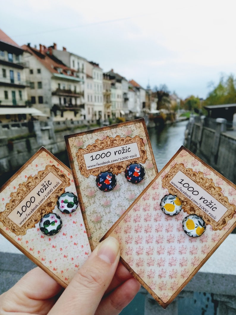 Puede incluir: Tres pares de pendientes en tarjetas con un patr&oacute;n floral. Los pendientes est&aacute;n hechos de vidrio y presentan diferentes dise&ntilde;os: un zorro rojo, un cactus y limones. Las tarjetas tienen el texto "1000 rozic" y una direcci&oacute;n de sitio web.