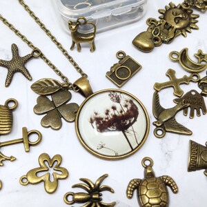May include: A collection of bronze-colored metal charms, including a four-leaf clover, a starfish, a bird, a cross, a crescent moon, a sun, a heart, a camera, a hand with the word "Hand Made" on it, a mushroom, a turtle, a bell, a spider, and a four-leaf clover with a leaf attached to it. The charms are arranged on a white surface. A necklace with a glass pendant featuring a dandelion is also included.