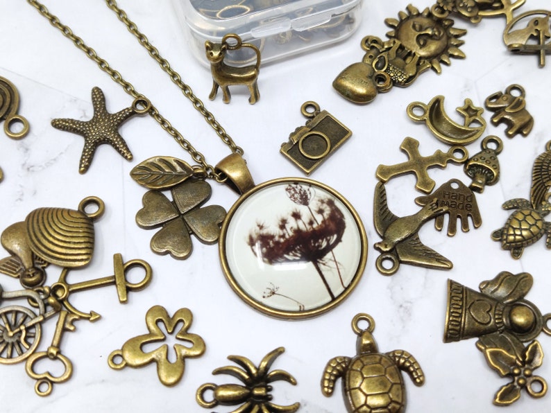 May include: A collection of bronze-colored metal charms, including a four-leaf clover, a starfish, a bird, a cross, a crescent moon, a sun, a heart, a camera, a hand with the word "Hand Made" on it, a mushroom, a turtle, a bell, a spider, and a four-leaf clover with a leaf attached to it. The charms are arranged on a white surface. A necklace with a glass pendant featuring a dandelion is also included.