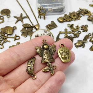 May include: Assortment of bronze-colored metal charms, including a skull, angel, and a mask. The charms are on a white surface.