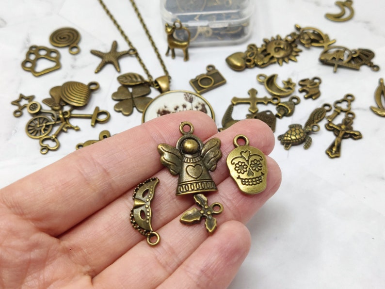 May include: Assortment of bronze-colored metal charms, including a skull, angel, and a mask. The charms are on a white surface.