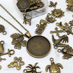 May include: A collection of antique bronze charms and pendants, including a four-leaf clover, a spiral, a turtle, a bird, a cross, a star, a mushroom, a camera, a deer, a heart, a sun, a  'hand made' tag, and a round pendant with a decorative design. The charms are arranged on a white surface.