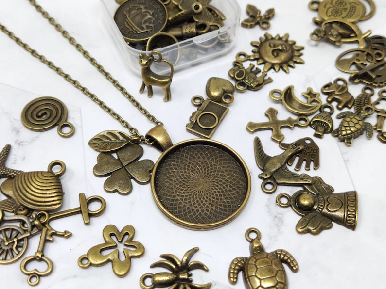 May include: A collection of antique bronze charms and pendants, including a four-leaf clover, a spiral, a turtle, a bird, a cross, a star, a mushroom, a camera, a deer, a heart, a sun, a  'hand made' tag, and a round pendant with a decorative design. The charms are arranged on a white surface.
