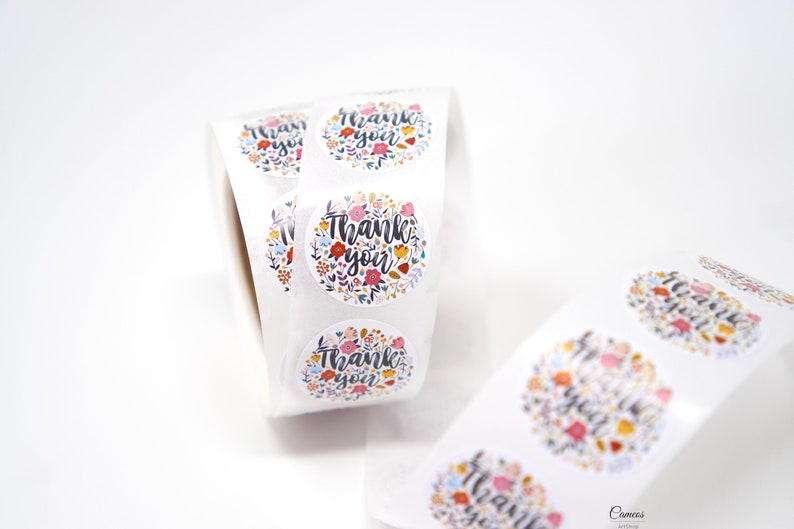 Pu&ograve; includere: Rotolo di adesivi rotondi con un design floreale e il testo "Grazie" in lettere nere. Gli adesivi sono bianchi con un motivo floreale colorato.