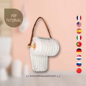 Puede incluir: Rollo de papel higiénico de crochet blanco con un asa de cuerda marrón. El rollo está colgado de una clavija de madera. El texto "La Fee Qui Pete" es visible en el rollo. El texto "www.lafeequipette.com" es visible en la parte inferior de la imagen.