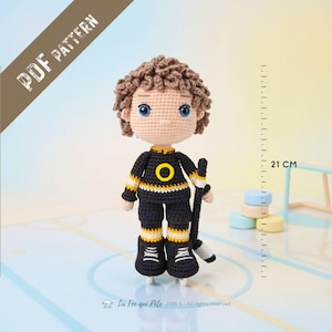 May include: A crochet doll of a hockey player with brown curly hair and blue eyes, wearing a black jersey with a yellow circle, black pants, and skates. The doll holds a hockey stick and stands 21 cm tall as indicated by a visible ruler.