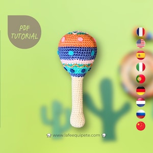 Puede incluir: Una maraca de crochet colorida con un mango blanco. La maraca tiene un patrón azul, rosa, naranja y verde. La maraca es un instrumento musical tradicional mexicano.
