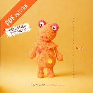 Peut inclure: Une peluche orange crochetée avec des yeux roses et blancs, un col à volants et deux taches colorées. Les textes "PDF PATTERN" et "BEGINNER FRIENDLY" sont visibles. Le jouet mesure 17 cm de haut.