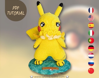 May include: A yellow crocheted Pikachu plush toy with a Cthulhu-inspired face. The toy is sitting on a blue and green crocheted base. The base has a label with the text "La Fee Qui Pique" and a website address.