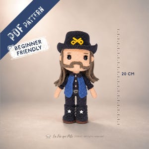 Peut inclure: Une poupée au crochet avec un chapeau noir orné d'un emblème jaune, de longs cheveux bruns, une moustache, un gilet bleu sur une chemise noire et un pantalon noir avec des étoiles blanches. Une règle indique 20 cm. Texte PDF PATTERN.