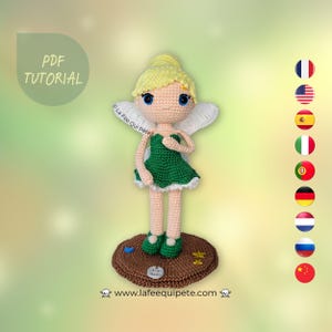 Könnte beinhalten: Häkelanleitung für eine grüne Feenpuppe mit blonden Haaren und Flügeln. Die Puppe steht auf einem braunen Sockel mit einem gelben Stern und einer blauen Blume. Das Muster ist als PDF-Download verfügbar. Der Text "PDF TUTORIAL" befindet sich in einem grünen Kreis oben im Bild. Der Text "La Fée Qui Pète" befindet sich in einem weißen Kreis unten im Bild. Der Text "www.lafeequipete.com" befindet sich unten im Bild.