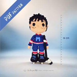 Peut inclure: Poupée de joueur de hockey au crochet faite à la main, d'environ 19 cm de haut, portant un uniforme bleu avec la lettre "M". La poupée a des cheveux noirs, des taches de rousseur et tient une crosse de hockey. Le texte "PDF PATTERN" est visible.