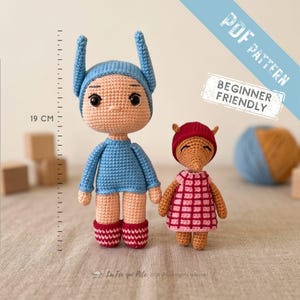Peut inclure: Deux poupées au crochet, une grande poupée bleue avec de hautes oreilles et une plus petite poupée beige avec un bonnet rouge et une robe à carreaux roses. Une règle indique 19 cm. Texte : PDF PATTERN BEGINNER FRIENDLY.
