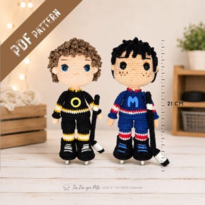 May include: Two crochet dolls standing 21 cm tall, each holding a hockey stick. One doll has brown curly hair and a black jersey with a yellow O, while the other has black hair and a blue jersey with a blue M. Text reads PDF PATTERN.