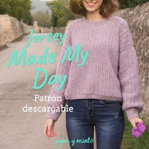 Puede incluir: Una mujer lleva un suéter de punto lavanda con el texto "Jersey Made My Day" y "Patrón descargable" en letras turquesa. El suéter es de corte holgado y tiene una textura acanalada. La mujer está de pie en un camino con un muro de piedra en el fondo.