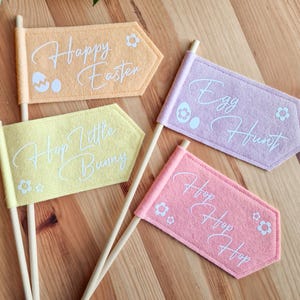 May include: Four felt Easter flags with white lettering and flower designs. The flags read "Happy Easter", "Hop Little Bunny", "Egg Hunt", and "Hop Hop Hop".