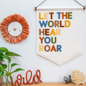 May include: A white felt banner with the text "Let the world hear you roar" in colourful felt letters. The banner is hanging on a white wall with a brown wooden dowel rod. There is a brown paper mache lion head, a red yarn word "wild", and a wooden leaf on a table below the banner.