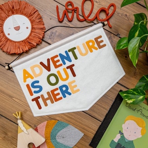 May include: A white felt banner with the words "Adventure is out there" in colorful felt letters. The banner is hanging on a wooden wall with other decorative items.