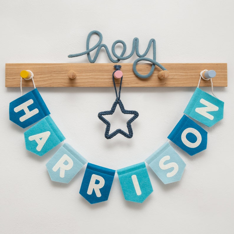 May include: A blue and white felt banner with the name "Harrison" spelled out in white letters. The banner is hanging from a wooden rail with a blue and white rope and a star-shaped decoration.