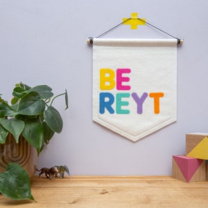 Peut inclure: Une bannière en feutre blanc avec les mots "BE REYT" en lettres de feutre arc-en-ciel. La bannière est accrochée à un mur violet clair avec un autocollant en forme de plus jaune au-dessus. Il y a trois blocs de bois au premier plan, peints en jaune, rose et bleu.