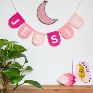 May include: A pink and white felt banner with the name "Jessie" spelled out in white letters. The banner is hanging from a white string and is decorated with a pink glass moon.