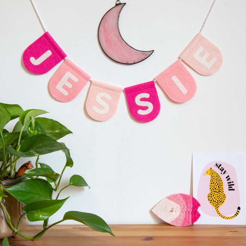 May include: A pink and white felt banner with the name "Jessie" spelled out in white letters. The banner is hanging from a white string and is decorated with a pink glass moon.