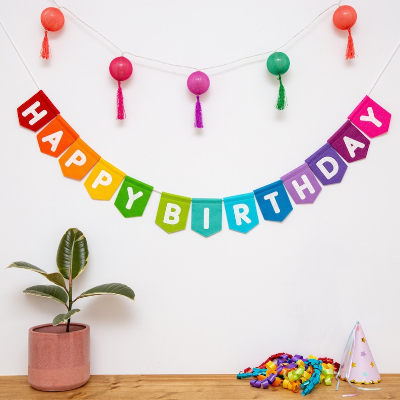 May include: A colorful felt banner spelling "HAPPY BIRTHDAY" hangs against a white wall, with a string of decorative lights and a potted plant. Party streamers and a party hat are on a wooden surface.