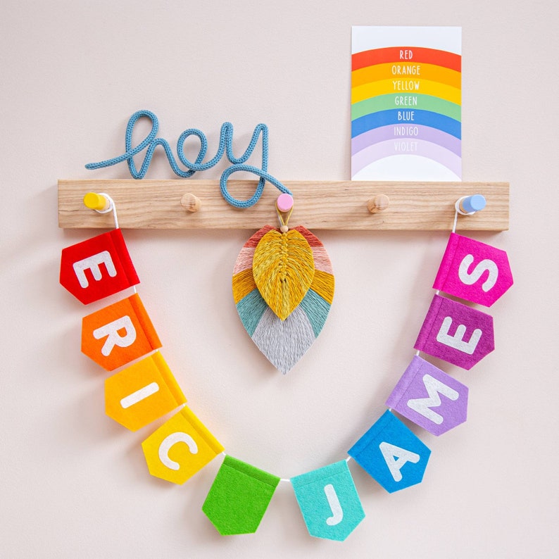 May include: A rainbow felt banner with the names Eric and James, with the word "hey" written in blue yarn above it. The banner is hanging on a wooden rack with hooks. A colorful feather decoration hangs in the center.