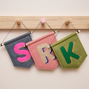 Könnte beinhalten: Drei Filz-Wimpel mit den Buchstaben "S", "R" und "K" in verschiedenen Farben und Stilen. Die Wimpel hängen an einer Holzstange.