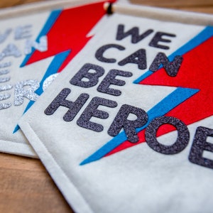May include: Two white felt banners with the text "We Can Be Heroes" in black glitter letters. The banners feature a red and blue lightning bolt design.