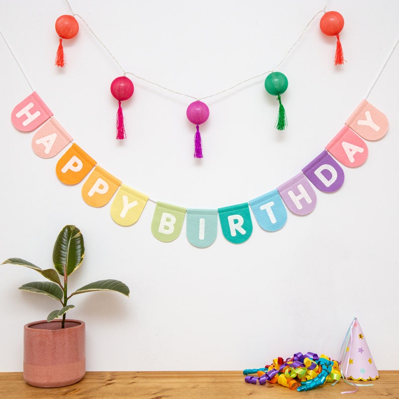 May include: A colorful "Happy Birthday" banner with white letters hangs against a white wall. Decorative string lights with red, pink, and green orbs and tassels are above the banner. A potted plant, party hat, and confetti are on a wooden surface.