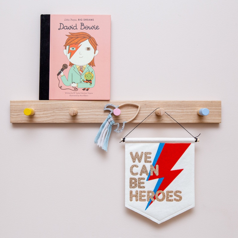 May include: A wooden wall-mounted rack with hooks, a book titled "Little People, Big Dreams: David Bowie", a blue and red banner with the text "We Can Be Heroes", and a blue and white tassel hanging from a hook.