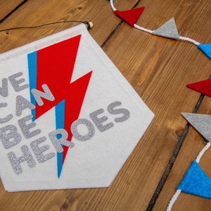 May include: A white felt banner with the text "WE CAN BE HEROES" in silver glitter letters. A red and blue lightning bolt design is behind the text. The banner is hanging from a wooden dowel rod with black string.